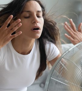 Eine junge Frau sitzt im Wohnzimmer vor einem Ventilator und versucht sich abzukühlen.