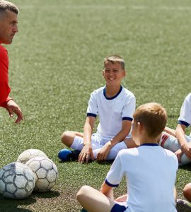 Eine Kinderfußballgruppe sitzt auf dem Rasen. Vor ihnen kniet ein Trainer. Vor dem Trainer liegt ein Fußball.